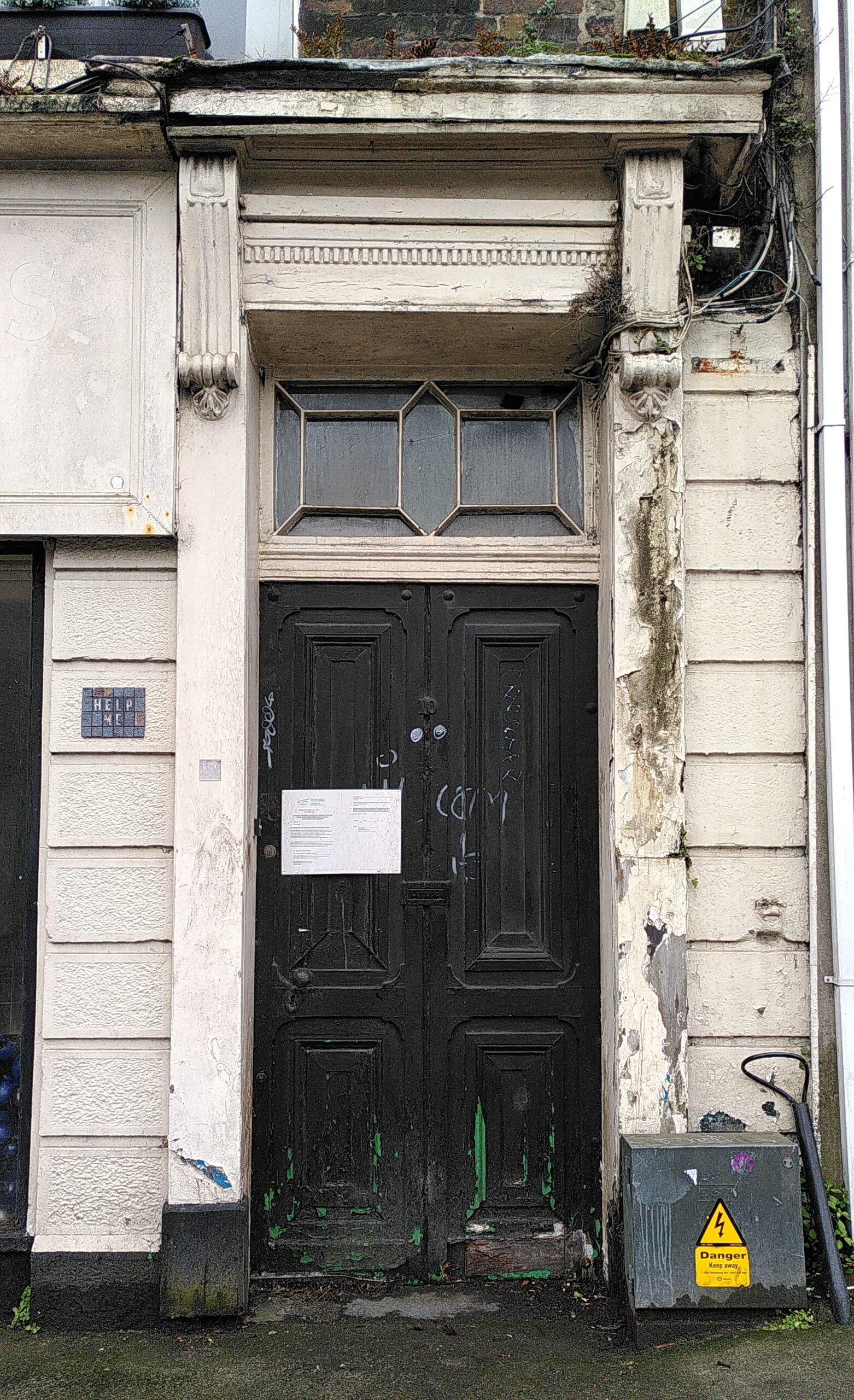 Door of the 10 Sarsfield Street - DS-033-23 (10/04/2025) Black wooden door, white decorated wooden frame with peeling paint. A notice on the door. On the left wall, a black mosaic tile.