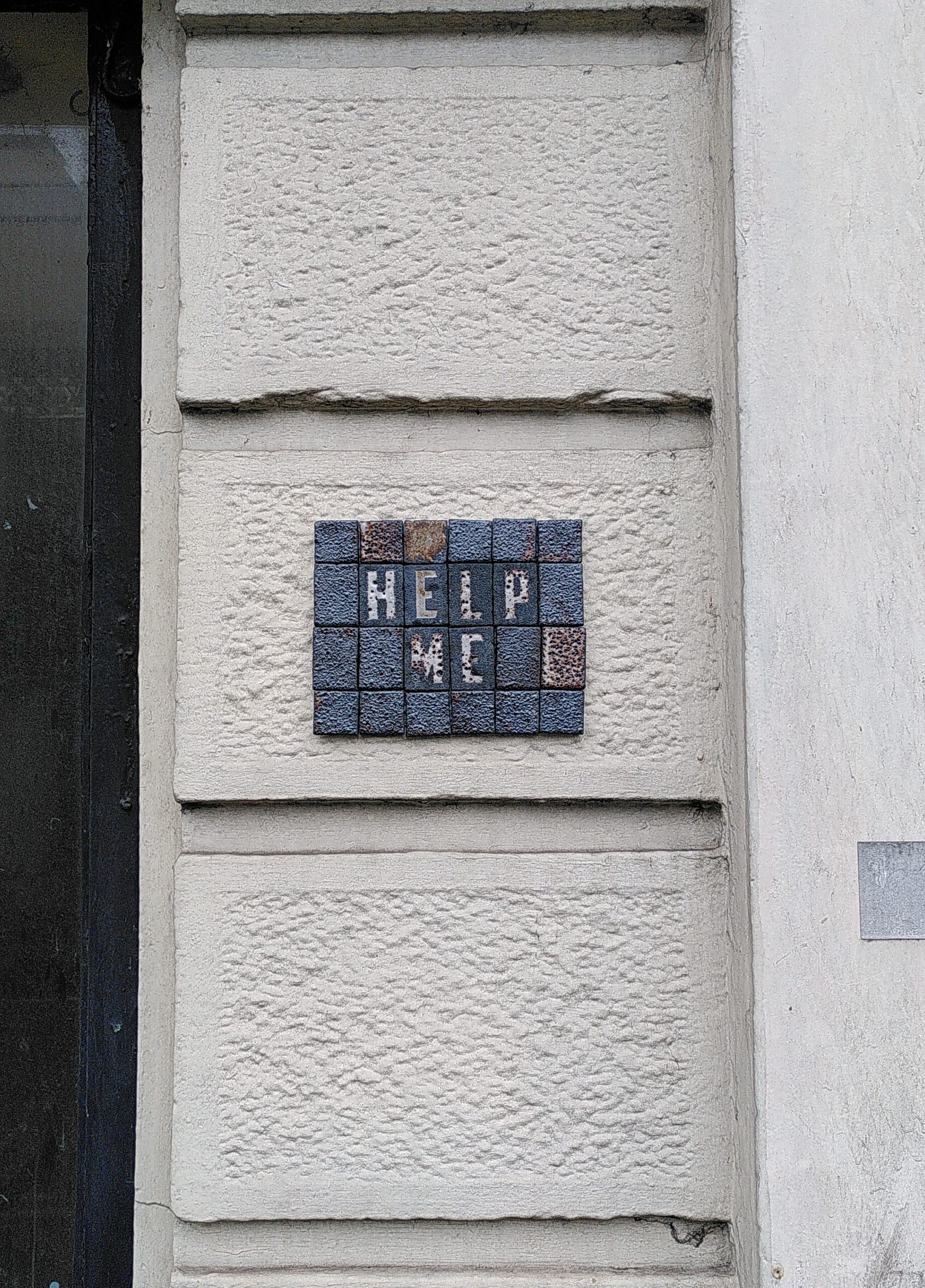 Mosaic tile "HELP ME" on wall of the 10 Sarsfield Street Ceramic mosaic tile. Rectangle. Black clay with a bubbly textured tiles. some glaze white/brown. Written HELP ME in capital letters. Glued on a white wall.