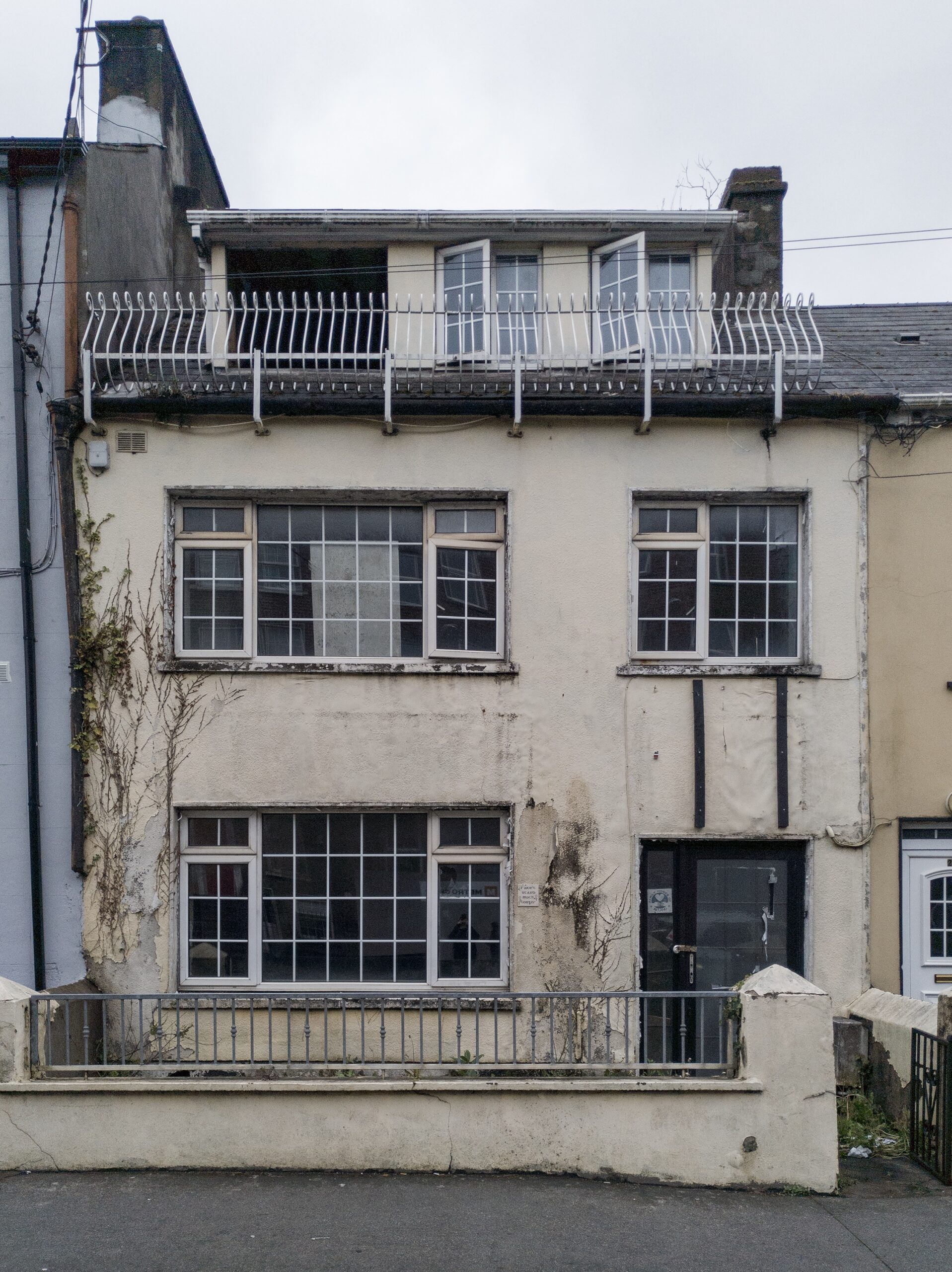 The front view of a beige house with dead plants growing on the facade here and there, some open or fully missing windows. Peeling paint. The house is looks like it's falling apart.