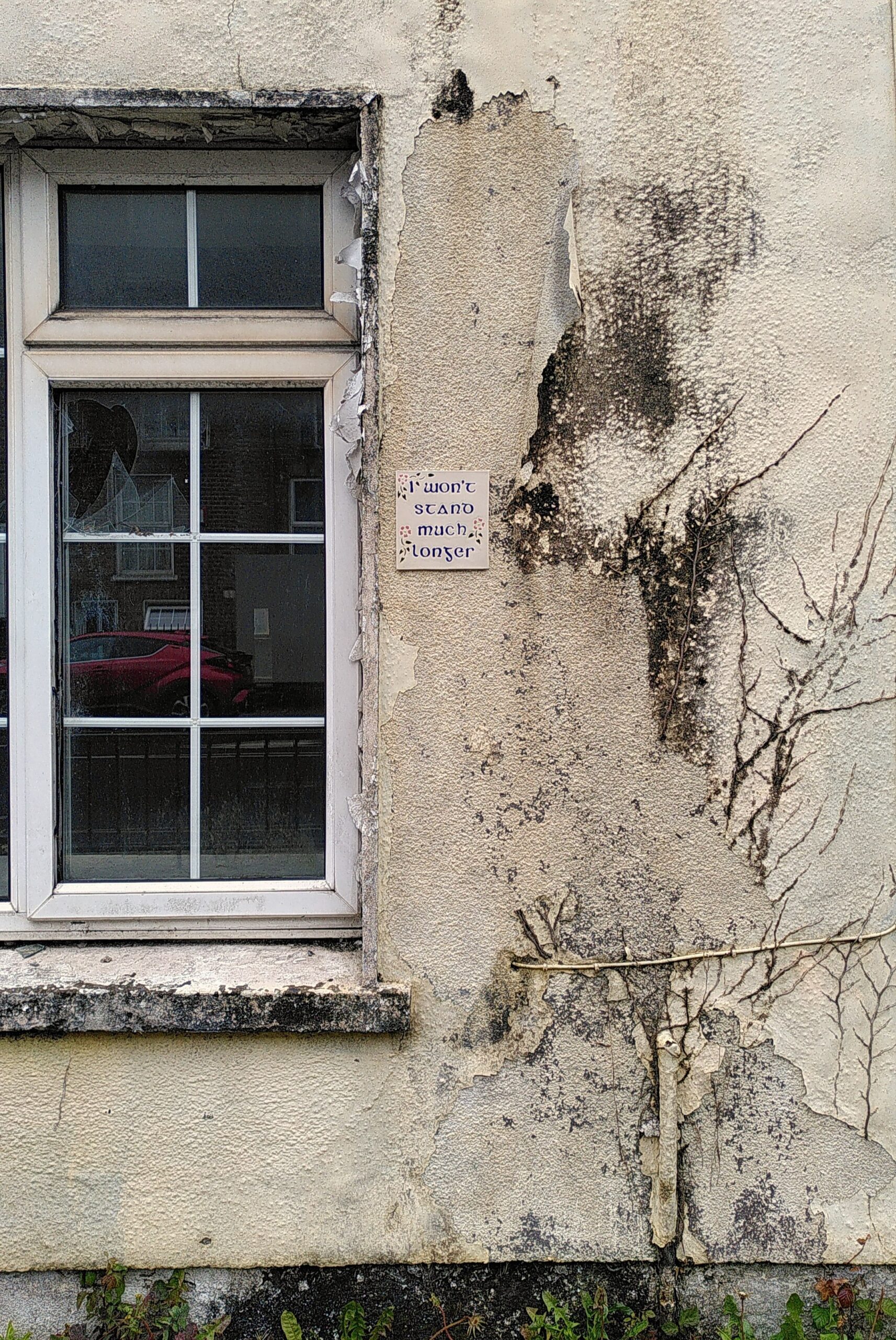 A part of a house wall. A part of window on the left. Paint is dirty and peeling off the wall in many places. Remnant of dead plants climbing up the wall. In the middle ceramic tile glued on the wall with written on it "I won't stand much longer".