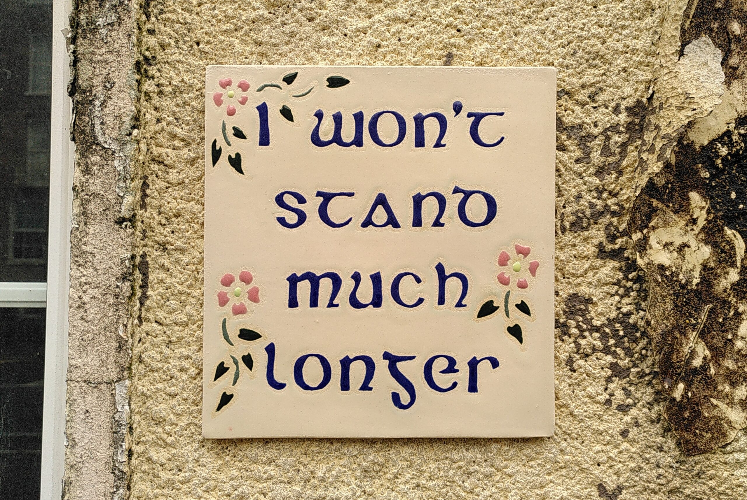 A ceramic tile written in blue Celtic style typo "I won't stand much longer" and decorated with pink little flowers and green leaves. Glued on a dirty beige wall with peeling paint.