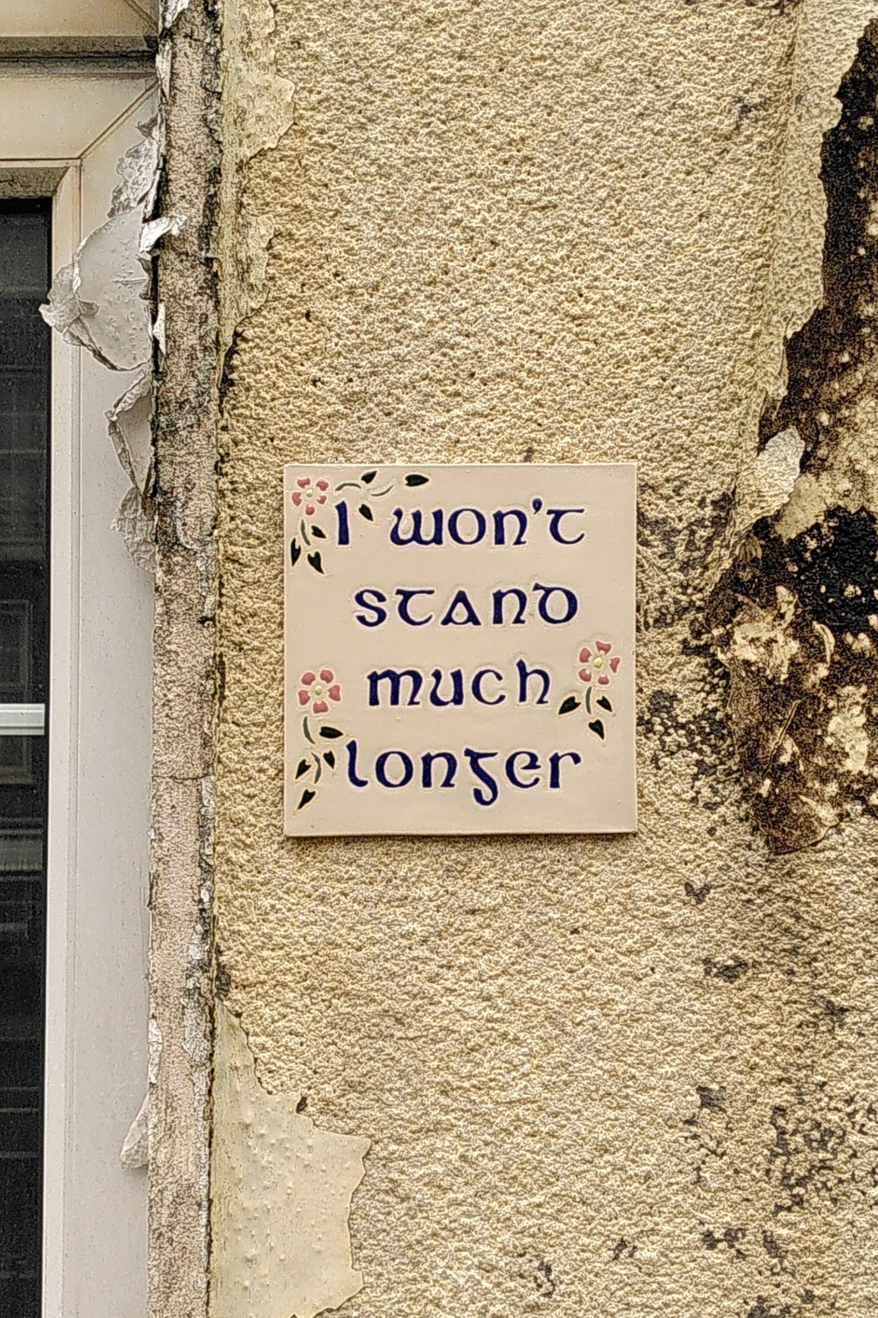 A part of a house wall. A part of window on the left. Paint is dirty and peeling off the wall. In the middle ceramic tile glued on the wall with written on it "I won't stand much longer" and decorated with small pink flowers and green leaves.