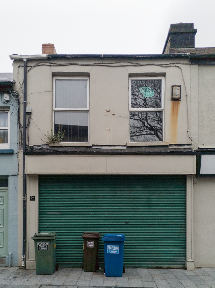 43-44 Nicholas street Front of a derelict building. Beige walls. Ground floor is a commercial unit and has green shutters closed. On 1st floor, big windows, plants growing on the left window.