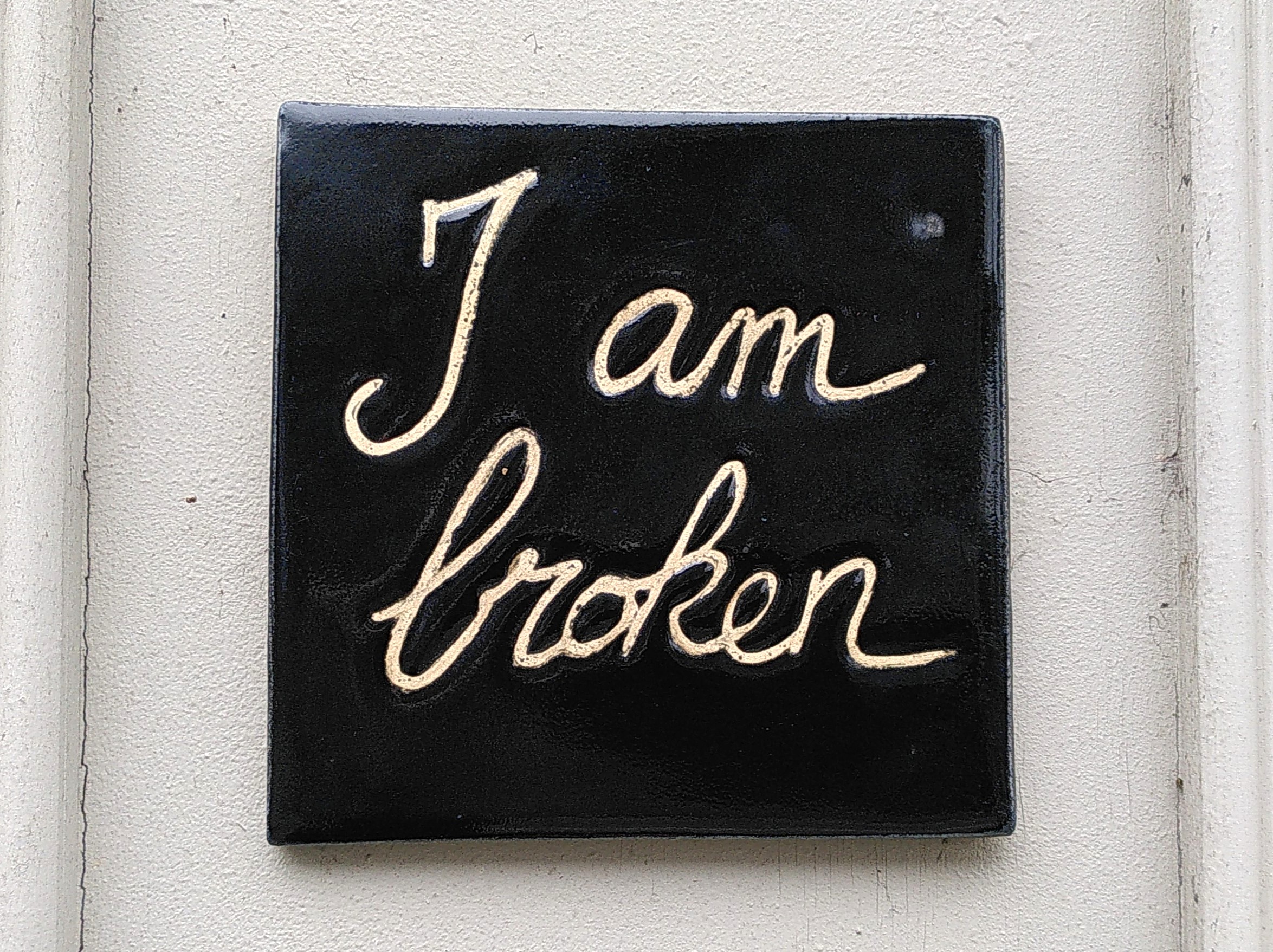 Ceramic tile "I am broken" A square ceramic tile. Black glaze. Message on it is handwritten "I am broken". It's glued on a white wall.