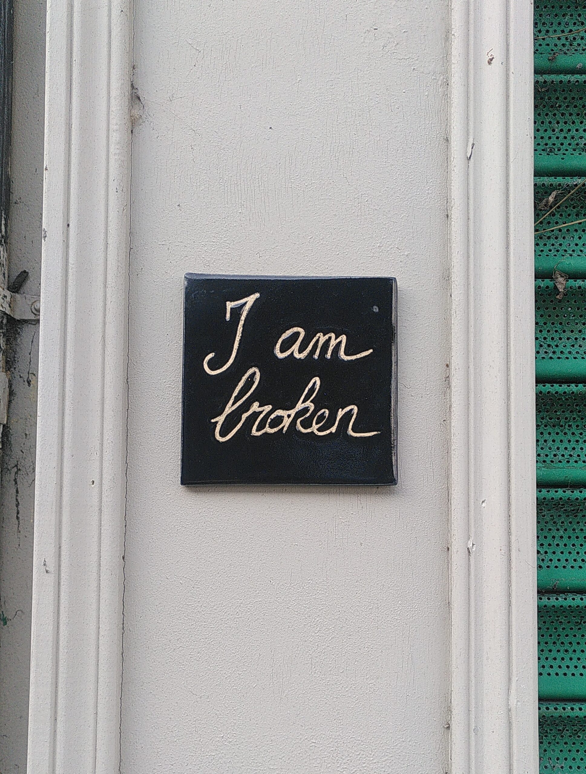 Ceramic tile "I am broken" A square ceramic tile. Black glaze. Message on it is handwritten "I am broken". It's glued on a white wall. On the right side of the picture we can start to see a green commercial shutter.