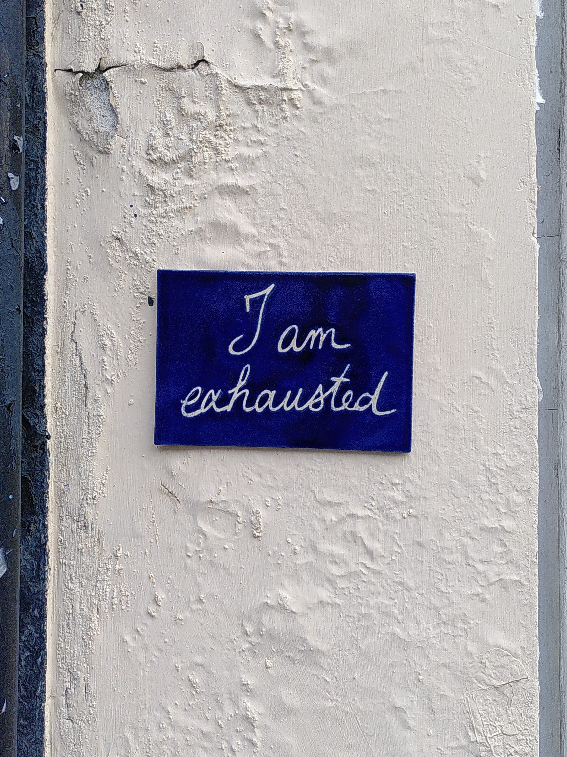 Ceramic tile "I am exhauted" A dark blue rectangle ceramic tile with handwritten in white "I am exhausted" on an off-white wall with peeling and bloating paint.