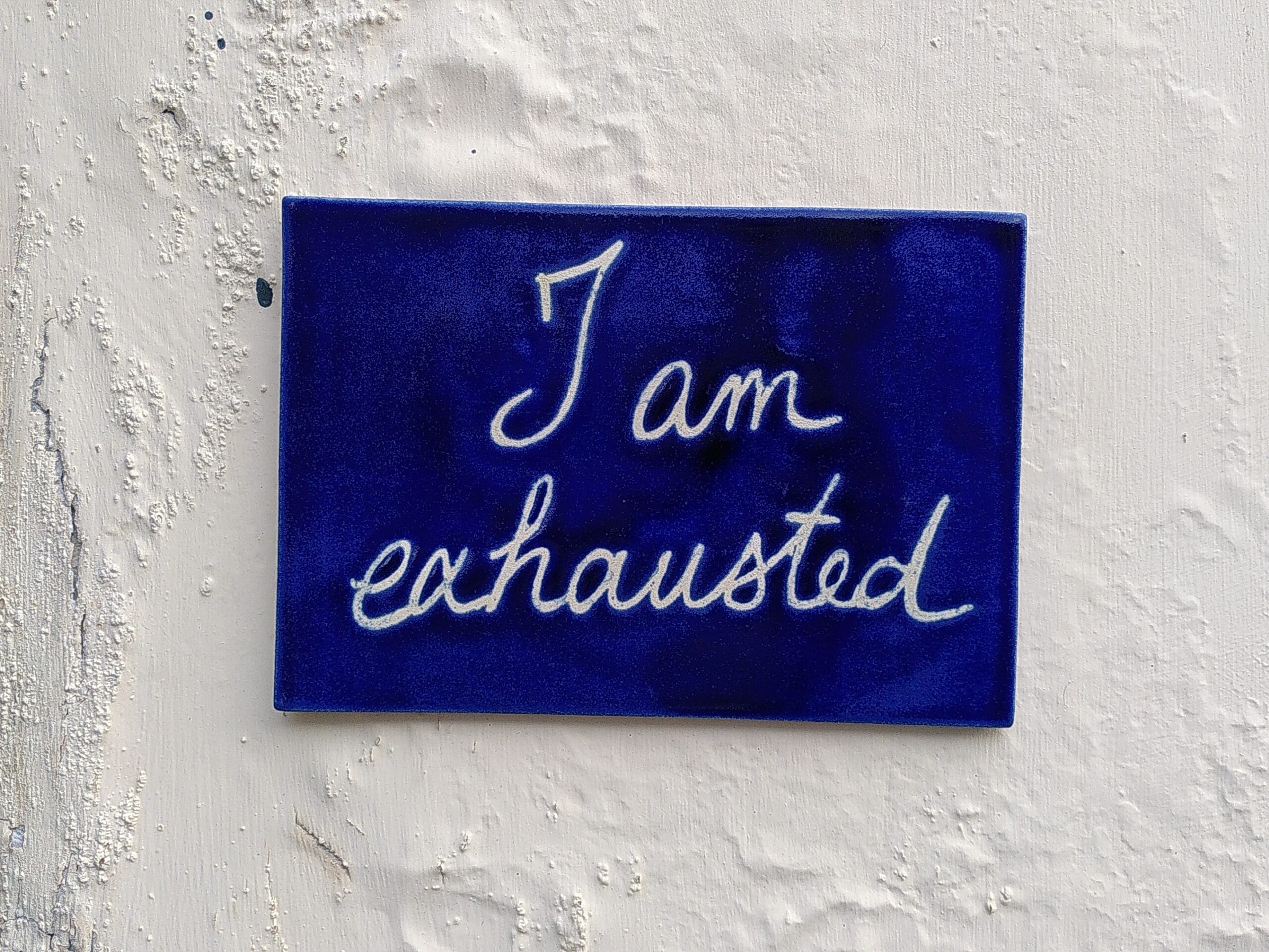 Ceramic tile "I am exhausted" A dark blue rectangle ceramic tile with handwritten in white "I am exhausted" on an off-white wall with peeling and bloating paint.