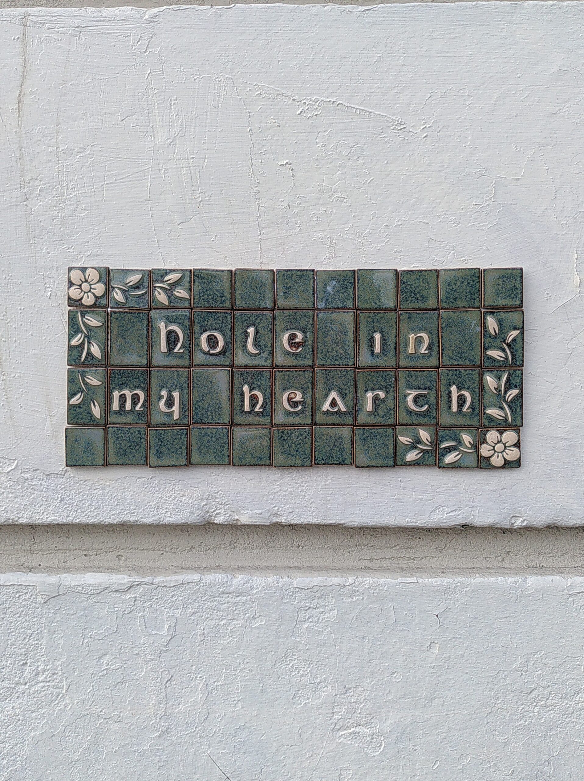 A ceramic mosaic plaque in a bleu green colour. Frame decoration are white flowers and leaves and text says Hole in my Hearth in white relief. The plaque is glued on a white wall.