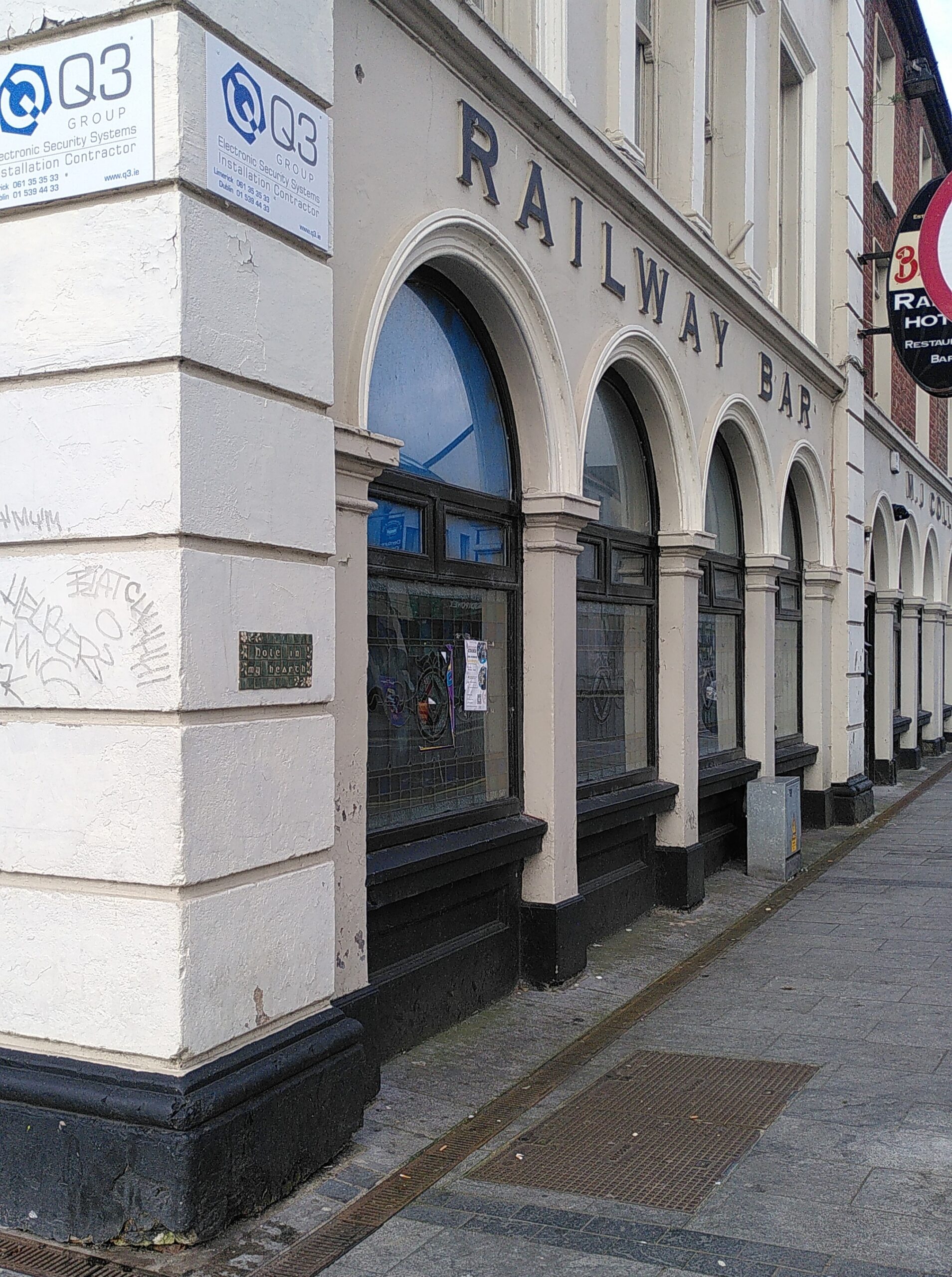 Photograph of the corner and side of the abandoned Railway Hotel in Limerick. Walls are white/cream. Arched windows with the name Railway Bar. On the corner we can see a ceramic plaque glued on the wall but we cannot see what is written on it.