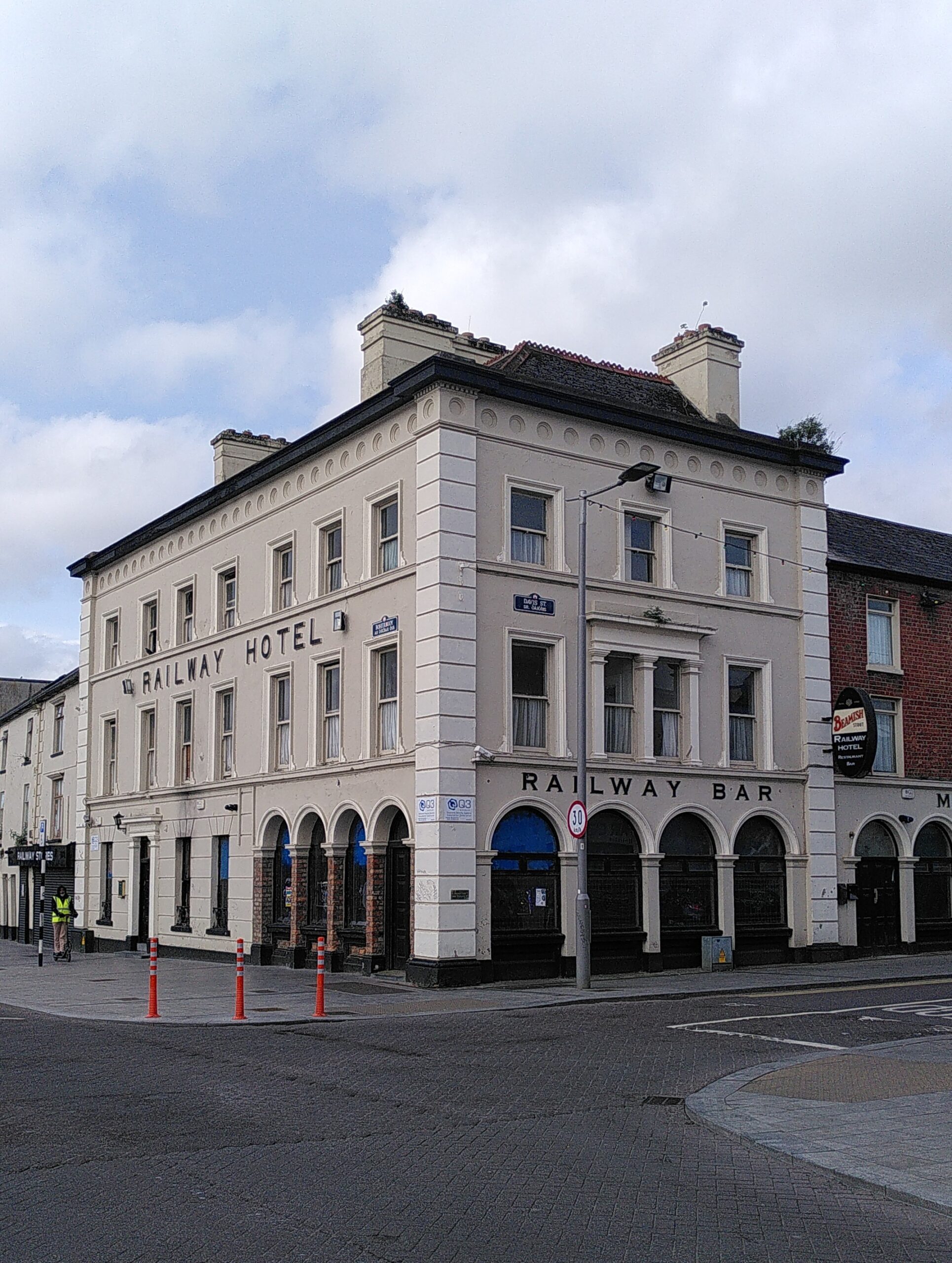 Railway hotel in Limerick city. It is a white decorated building with the name Railway Hotel in big capital letters on it. the ground part is the Railway Bar with arched windows. We can see plants growing everywhere on the roof. And guess some broken windows.