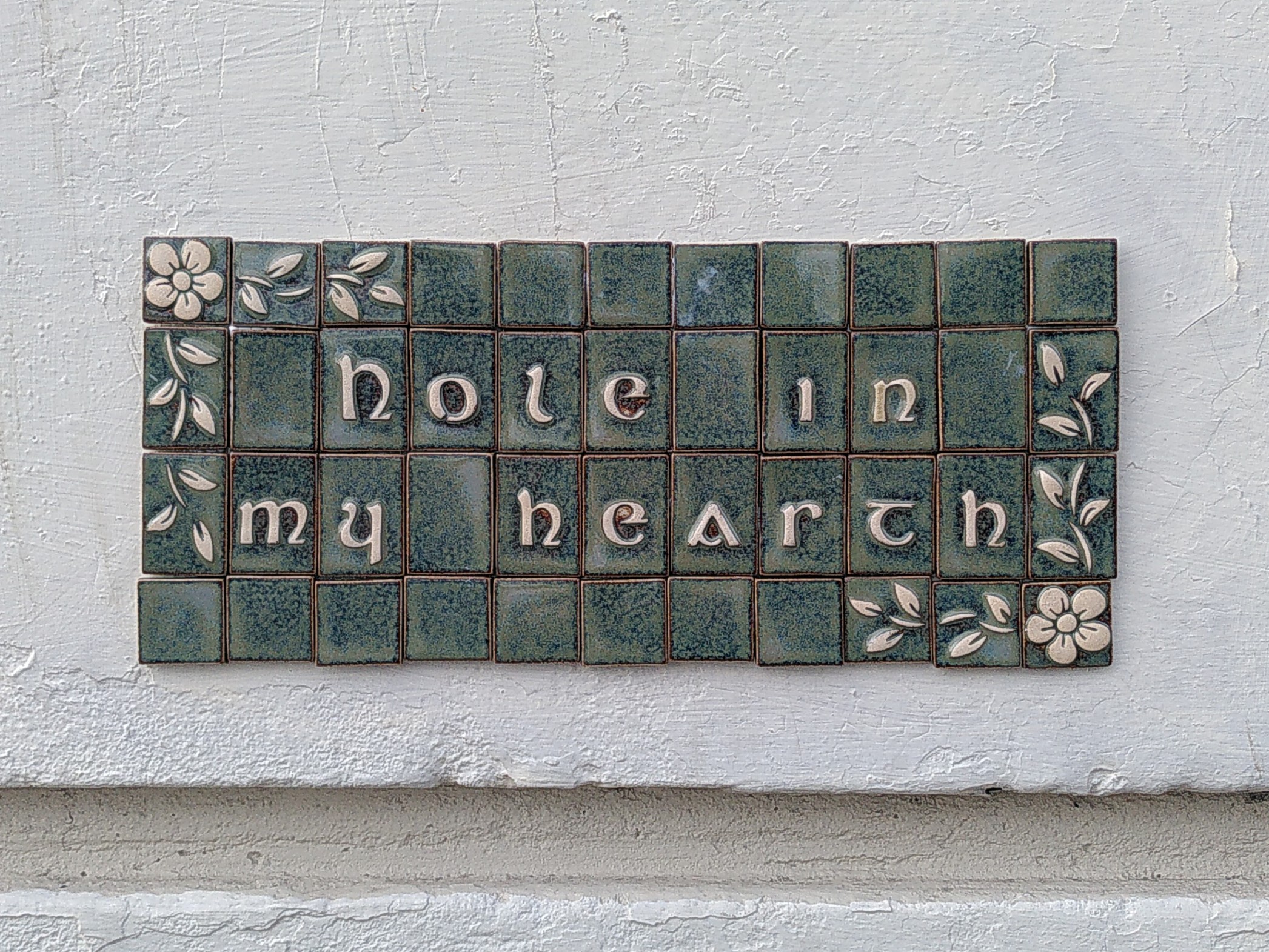 A ceramic mosaic plaque in a bleu green colour. Frame decoration are white flowers and leaves and text says Hole in my Hearth in white relief. The plaque is glued on a white wall.